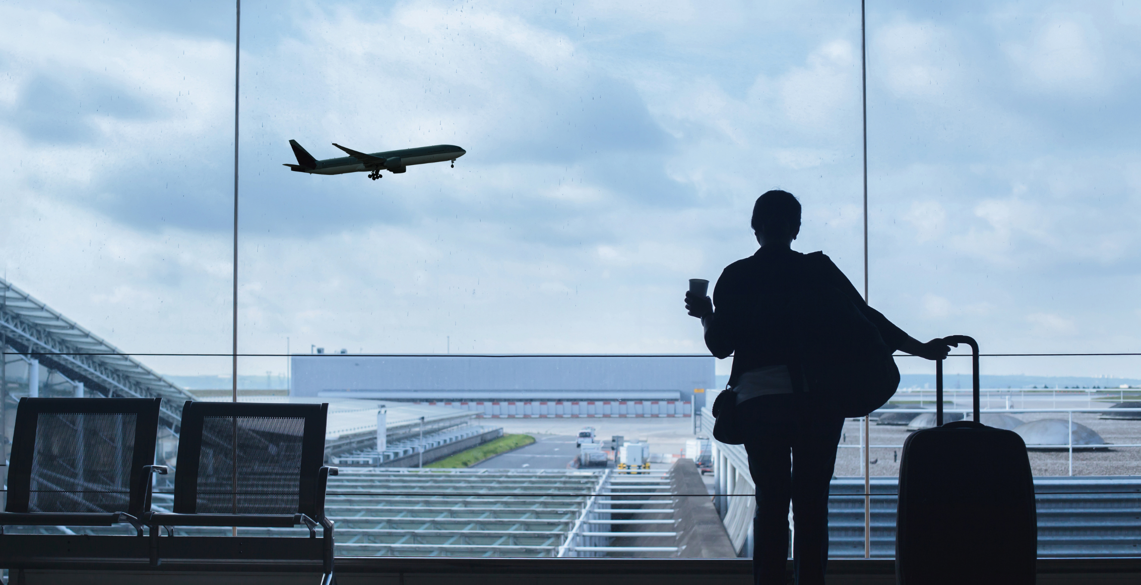 Person holds on to suitcase in airport and stares out the window as a plan takes off